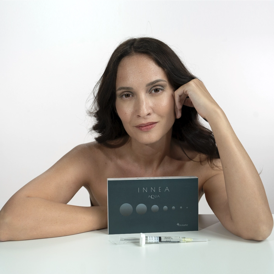 Confident woman presenting the INNEA Aqua product box, with injectable syringe displayed on the table. Highlights hydration and skin vitality through aesthetic innovation.