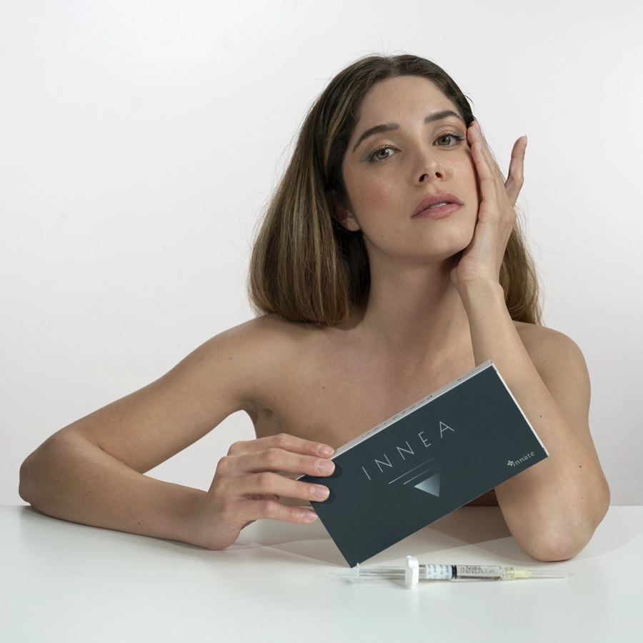 Young blonde woman with radiant skin holding an INNEA Classic product box, with injectable syringes displayed on the table. Represents confidence and precision in aesthetic treatments.