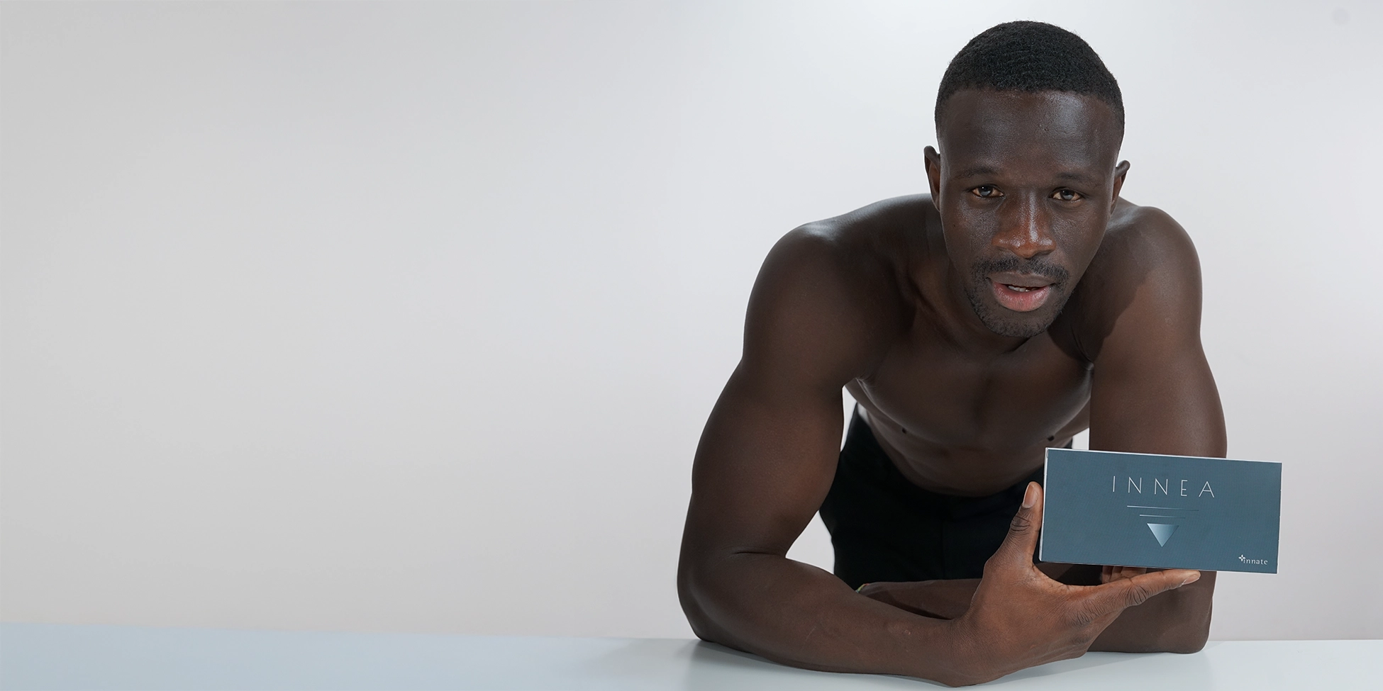 Man with defined physique leaning on a table, holding the INNEA Classic product box. Embodies strength and self-care in aesthetic medicine.